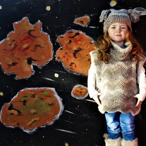 May include: A young girl wearing a gray crocheted vest with a white crocheted hat and blue jeans. The background is a black canvas with a painted space scene.