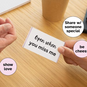 Pode incluir: Um cartão branco com o texto "Open when you miss me" é segurado entre duas mãos. O cartão tem uma borda roxa. A imagem também inclui as frases "show love", "Share w/ someone special" e "be cheesy" em círculos rosa.