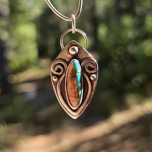 Peut inclure: Un collier pendentif en argent avec un pendentif en forme de goutte d'eau. Le pendentif présente une pierre brune centrale avec une bande bleu turquoise, encadrée par un sertissage argenté avec des volutes décoratives et de petites perles d'argent. Le collier est suspendu sur un fond vert flou.