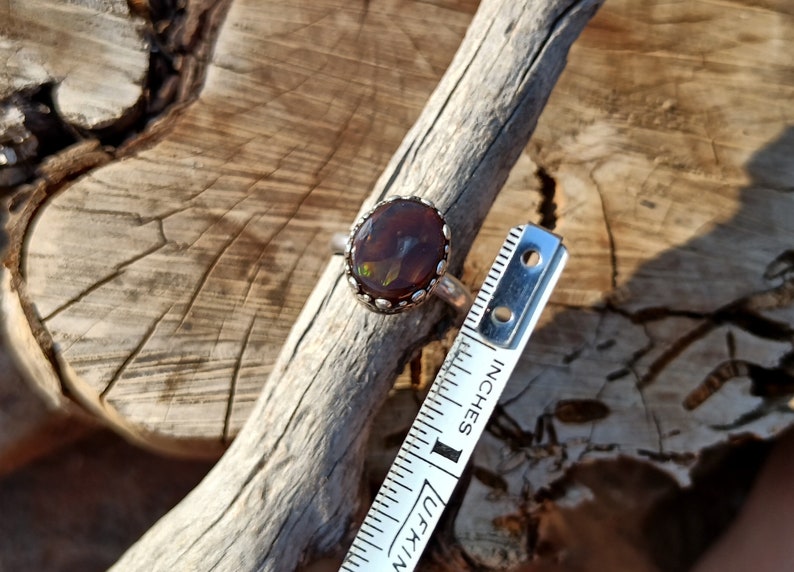 May include: A silver ring with a brown and green opal gemstone set in a silver bezel. The ring is shown next to a ruler measuring 1 inch.