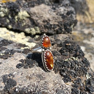 May include: A silver ring featuring two orange agate stones set in an ornate silver frame. The ring is resting on a rocky surface.