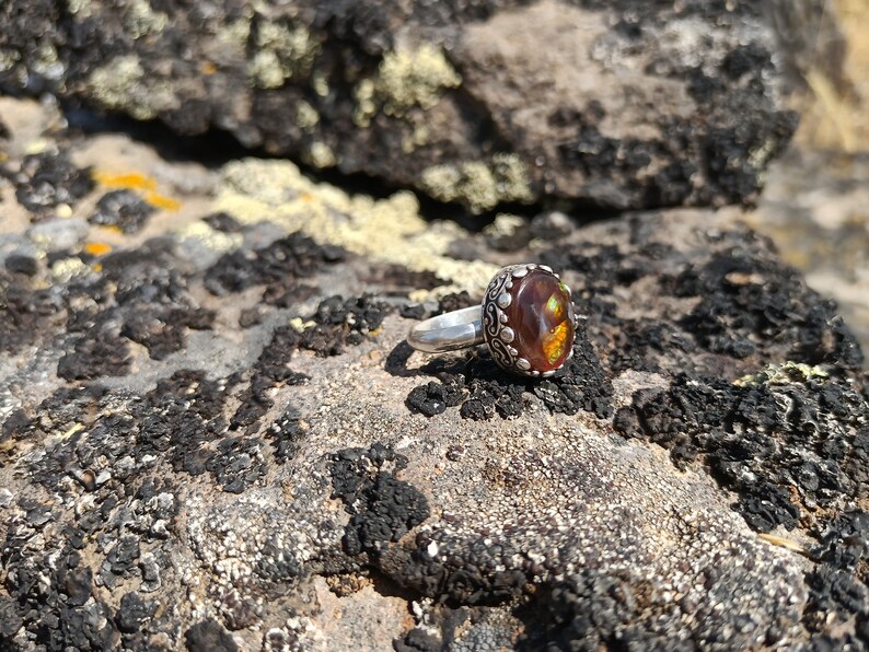 May include: A silver ring with a brown and gold gemstone set in a decorative silver bezel. The ring is resting on a rocky surface.