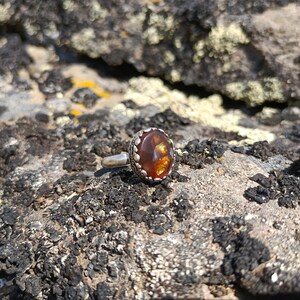 May include: A silver ring with a round, orange and green gemstone set in a silver bezel. The ring is on a rough, brown and gray rock.
