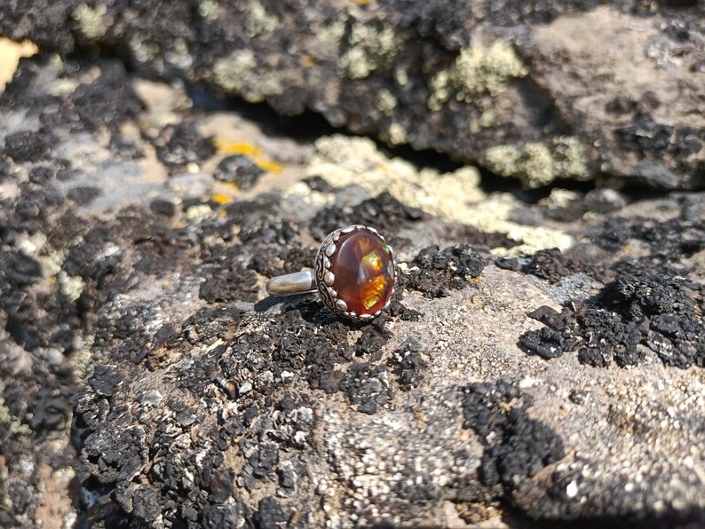 May include: A silver ring with a round, orange and green gemstone set in a silver bezel. The ring is on a rough, brown and gray rock.