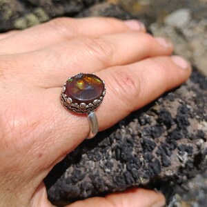 May include: A silver ring with a large, round, brown agate gemstone set in a decorative silver bezel. The ring is on a hand, resting on a rough, dark brown rock.