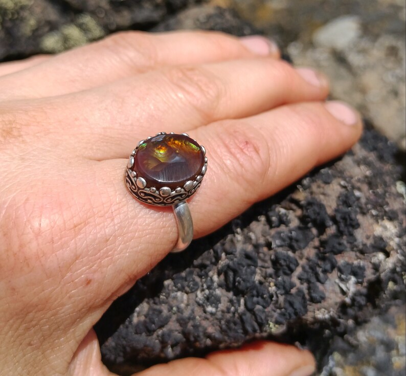 May include: A silver ring with a large, round, brown agate gemstone set in a decorative silver bezel. The ring is on a hand, resting on a rough, dark brown rock.