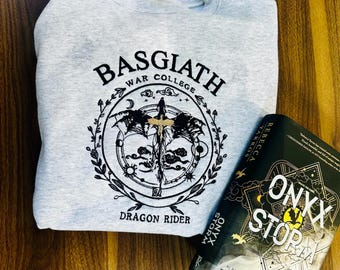 May include: A light gray sweatshirt with the text "BASGIATH WAR COLLEGE DRAGON RIDER" in black. The sweatshirt features a circular graphic with a sword and dragon wings. A book titled "ONYX STORM" by Rebecca Yarros is next to the sweatshirt.