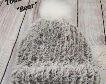 May include: A faux fur toddler hat in shades of grey and brown, with a large white pom-pom. The hat has a soft, textured appearance. The text "Toddler hat Bear" is visible on the image.