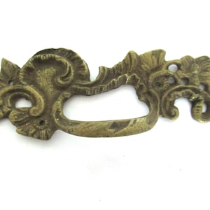 Może przedstawiać: Ozdobny mosiężny uchwyt do szuflady z zakrzywionym uchwytem i dekoracyjnym wzorem kwiatowym i przewijanym. Okucia w stylu antycznym mają ciepły, postarzany złoty kolor, idealny do renowacji mebli w stylu vintage.