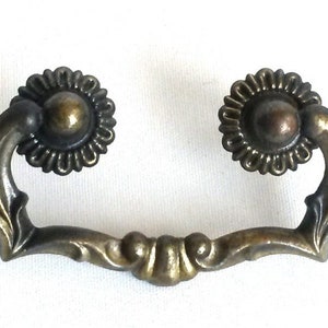 Könnte beinhalten: Ein antiker Messing-Schubladengriff mit floralem Design. Der Griff hat zwei verzierte Knöpfe mit einer erhabenen Mitte und eine gebogene Stange, die sie verbindet.