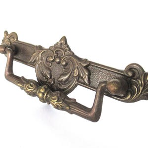 Può includere: Una maniglia per cassetto in ottone in stile vintage con un design ornamentale. La maniglia ha una maniglia curva e una piastra decorativa con intricati lavori a scorrimento.