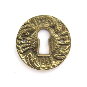 May include: A round, ornate brass keyhole cover with a detailed design. The cover is a solid piece of metal with a keyhole cut out in the centre.
