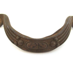 Op de afbeelding: Een rustieke, bruine metalen deurknop met een decoratief ontwerp. De knop heeft een gebogen vorm met een verhoogde ovale vorm in het midden en een reeks kleine, ronde accenten.