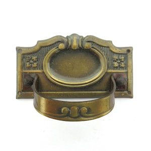 Puede incluir: Un tirador de cajón de latón vintage con un diseño ornamentado. El tirador tiene una base rectangular con un óvalo decorativo en el centro y un asa curva.