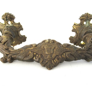 Peut inclure: Poignée de tiroir en laiton ornée avec un motif de lyre au centre. La poignée a un motif enroulé de chaque côté et une rosette décorative à chaque extrémité.