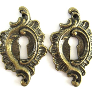 May include: Two ornate keyhole escutcheons in an antique brass finish. The escutcheons are shaped like a stylized flower with a keyhole in the centre.