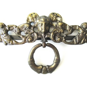 Könnte beinhalten: Antiker Schubladengriff aus Messing mit detailliertem Gesicht und Ringgriff. Die dekorative Hardware weist auf beiden Seiten des Gesichts kunstvolle Schnörkel und Figuren auf. Ein Akzent im Vintage-Stil für Möbel.