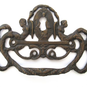 Op de afbeelding: Oornamentale bronzen ladeknop met een sleutelgatontwerp. De knop heeft een centraal sleutelgat, geflankeerd door twee cherubijnfiguren en decoratief krulwerk. De handgreep is gebogen en gedetailleerd, geschikt voor antieke meubels.