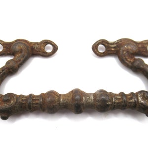 Puede incluir: Un tirador de cajón de hierro fundido oxidado con detalles ornamentados y una sección central decorativa. El tirador tiene dos agujeros para el montaje.