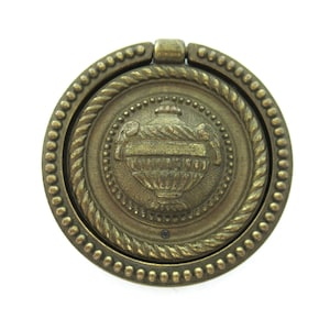 Könnte beinhalten: Ein runder, antiker goldfarbener Schrankknopf mit einem seilartigen Design am Rand und einem erhabenen, verzierten Urnen-Design in der Mitte.