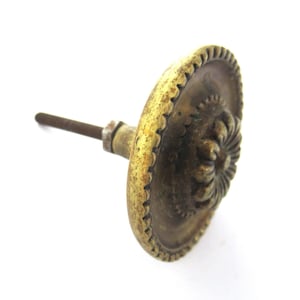 以下が含まれることがあります： 装飾的な花柄とギザギザの縁を持つヴィンテージの真鍮製のドアノブ。ノブは摩耗しており、パティナ仕上げになっています。