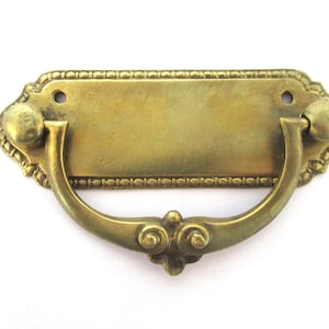 Puede incluir: Un tirador de cajón de latón con una placa rectangular y una manija decorativa. La placa tiene un borde con cuentas y la manija tiene un diseño de desplazamiento.