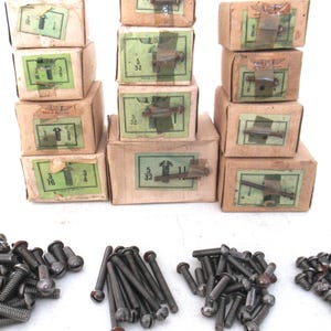Peut inclure: Une collection de boîtes de vis vintage, avec une variété de tailles et de finitions. Les boîtes sont en carton et ont un schéma de couleurs brun et vert. Les vis sont en métal et ont une finition noire. Les boîtes sont empilées les unes sur les autres, et il y a plusieurs piles de vis éparpillées autour des boîtes.