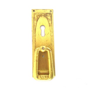Könnte beinhalten: Ein goldfarbener Türgriff mit einem dekorativen Design und einem Schlüsselloch. Der Griff ist an einer rechteckigen Platte mit einem erhöhten Rand befestigt.