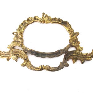 Puede incluir: Tirador de cajón de latón antiguo y ornamentado, con un centro ovalado abierto y detalles de volutas. El herraje tiene un aspecto deslustrado y envejecido, lo que sugiere un uso vintage o reutilizado. Ideal para la restauración de muebles.