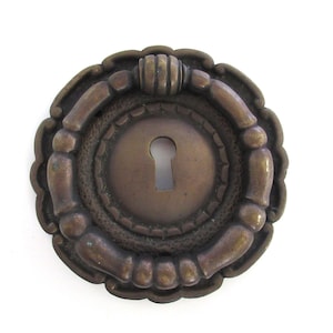 May include: A round, ornate brass keyhole cover with a decorative ring around the keyhole. The cover has a raised, textured design and a smooth, polished finish.