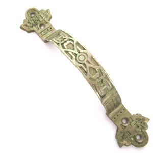 Può includere: Una maniglia per cassetto in ottone antico con un design curvo e ornato. La maniglia presenta un motivo traforato dettagliato ed estremità decorative con fori di montaggio. L'ottone ha una patina verdastra leggermente invecchiata.