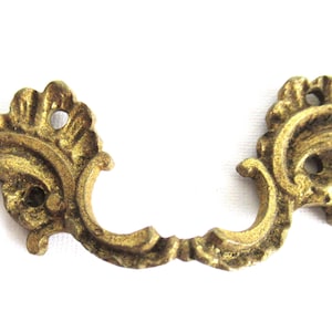 Op de afbeelding: Een paar sierlijke gouden metalen ladegrepen met een decoratief bloemmotief. De grepen zijn C-vormig en hebben elk drie gaten.