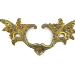 May include: A gold-toned metal decorative drawer pull with a floral design. The pull has two ornate wings on each side and a curved handle in the centre.
