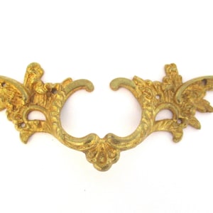 Peut inclure: Poignée de tiroir dorée ornée avec un design symétrique. La poignée présente une zone de poignée centrale avec des ornements décoratifs et deux éléments en forme d'ailes de chaque côté. Pour meubles anciens.