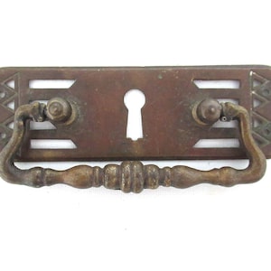 May include: A brass drawer pull with a keyhole cutout and an ornate design. The pull has a rectangular base and a curved handle.