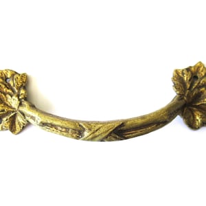 Może przedstawiać: Złoty metalowy klamka do drzwi z wzorem winorośli. Klamka ma zakrzywiony kształt i jest ozdobiona liśćmi i winogronami.