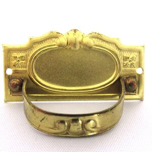 Può includere: Una maniglia per cassetto in metallo dorato con una piastra ovale e una maniglia decorativa. La piastra ha un bordo rialzato e un motivo floreale.