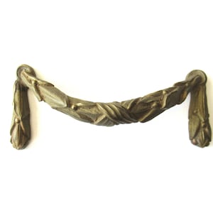 Puede incluir: Un tirador de armario de metal dorado con un diseño decorativo retorcido. El tirador tiene una forma curva y un acabado liso.