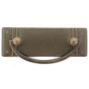 Può includere: Una maniglia per cassetto rettangolare in metallo marrone con una maniglia curva e due pomelli rotondi.