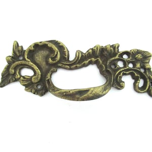 Könnte beinhalten: Verzierter antiker Schubladengriff aus Messing mit gebogenem Griff und dekorativem Blumen- und Schnörkeldesign. Die Hardware hat eine warme, gealterte Messingfarbe, ideal für Restaurierungsprojekte von Vintage-Möbeln.