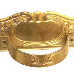 Könnte beinhalten: Ein goldfarbener Türgriff aus Metall mit einem verzierten Design. Der Griff hat eine rechteckige Basis mit einer gewölbten Oberseite und einem ringförmigen Griff.