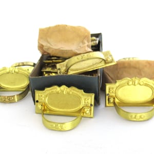 Peut inclure: Une boîte de tirettes de tiroir en laiton vintage avec des motifs ornés. Les tirettes ont une plaque rectangulaire avec une découpe ovale et une poignée incurvée. La boîte est remplie de nombreuses tirettes et est partiellement ouverte, révélant les tirettes à l'intérieur.