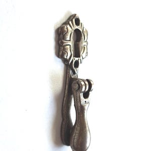 May include: A metallic, antique-toned furniture pull featuring a decorative, scalloped escutcheon plate with a central keyhole opening. The hardware includes a hinged, teardrop-shaped drop handle that hangs vertically from a central pivot point.