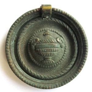 Peut inclure: Bouton de tiroir antique de couleur bronze. Le centre présente un motif d'urne détaillé dans un cercle de style corde. Une petite bélière rectangulaire de couleur laiton est fixée en haut. Le bouton de tiroir a un aspect vieilli.