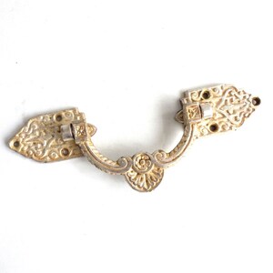 May include: A vintage drawer pull in a tarnished gold finish, featuring intricate floral and scrollwork designs. The handle curves gracefully, with decorative arrowhead-shaped ends. The metal hardware is designed for furniture restoration.