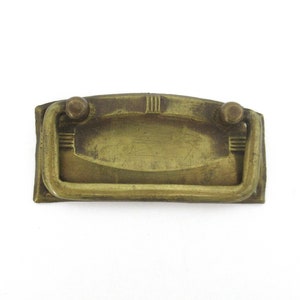 Può includere: Una maniglia per cassetto in ottone con forma rettangolare e bordi arrotondati. La maniglia ha un design semplice con una sezione centrale rialzata e due piccoli pomelli su entrambi i lati.