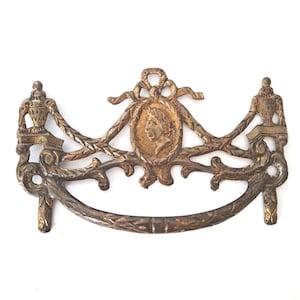 May include: A brass drawer pull featuring a central oval medallion with a profile portrait of a person. The hardware includes a curved handle, decorative swags, and two urn-shaped finials on the sides, all finished with a weathered patina.