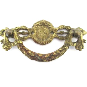 Op de afbeelding: Een gouden metalen ladegreep met een decoratief, sierlijk ontwerp. De greep heeft een centrale ovale vorm met een reliëf, gestructureerd oppervlak en ingewikkelde bloemmotieven.