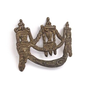 Op de afbeelding: Oud bronzen ladeknop met een sierlijk ontwerp. De knop heeft een centraal vaasachtig element geflankeerd door twee verticale, gestileerde figuren, verbonden door gebogen armen. Het brons heeft een verweerde uitstraling.