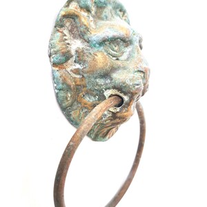 Op de afbeelding: Een antieke bronzen deurklopper met een leeuwenkop en een ringhandvat. De leeuwenkop heeft een verweerde uitstraling met groene en bruine patina. De ringhandgreep heeft een roestbruine kleur.
